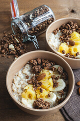 Oatmeal with pineapple, banana and chocolate granola in beige plates on a brown wooden table. A hearty healthy breakfast. A place for text.