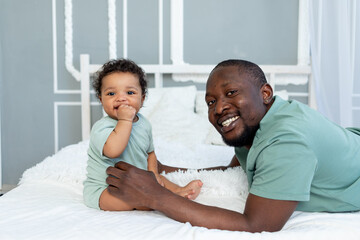 happy smiling African american dad with baby son on bed at home cuddling and playing, happy family, father's day