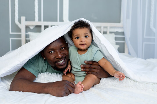 happy smiling African american dad with baby son on the bed at home cuddling under the blanket, happy family, father's day