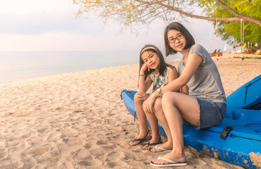 Fototapeta premium Beautiful Asian mother and child sit on kayak boat, pose on the sandy beach, blurred background of sea and sky, perspective view, warm tone image with blank space for copy and design.