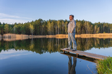 Fototapeta na wymiar Man stands on a wooden pier near spring forest on a calm lake in Ukraine. Nature and travel concept