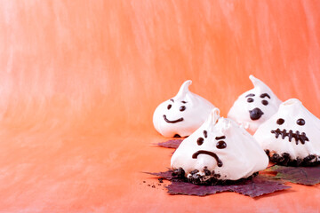 Funny meringue ghosts with funny faces against the orange background. Halloween dessert