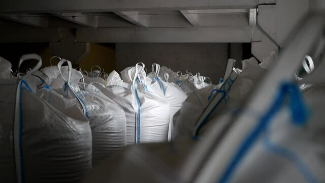 Close-up large, full bags of cereal products in stock. The bags are stacked in rows, industrial warehouse. Big white plastic bags with cereals: rye, wheat, barley, mustard are storing in storage.