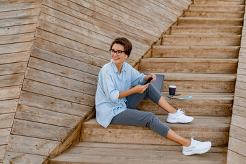 young pretty woman sitting with laptop in modern park street using smartphone