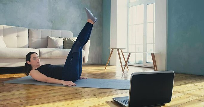 Active young woman in skinny tracksuit does crunches and leg lifts watching online lesson on laptop at home training slow motion