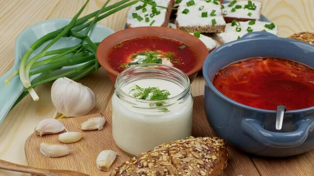 Russian and Ukrainian cuisine. Borscht with sour cream and spices, fresh herbs, garlic and pampushki. Beetroot soup in a blue bowl on a wooden board.