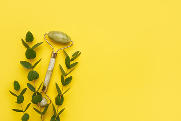 Green face roller from natural jade stone and eucalyptus branches on a yellow background. SPA concept