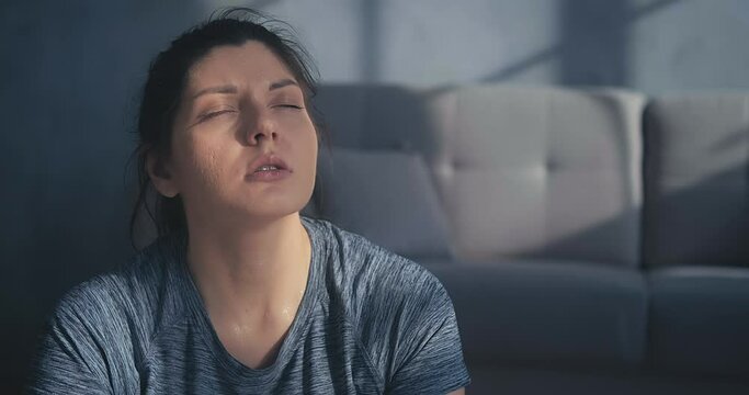 Tired young woman in grey t-shirt lit by sunlight rests breathing heavily after intensive training in room closeup slow motion