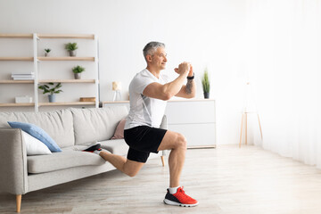 Active mature man doing squats while training at home, doing exercises near sofa in living room...