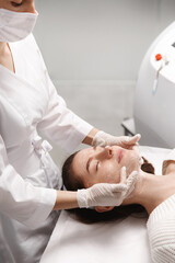 Beautician in beauty clinic smear cleansing skincare product onto woman skin to gently clean and nourish facial cells before ELOS hardware cosmetology treatment