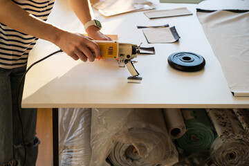 Closeup shot of professional female tailor using electric cutter to cut clothing design samples pattern on table in production studio. Working in tailoring shop concept. Busy woman dressmaker at work