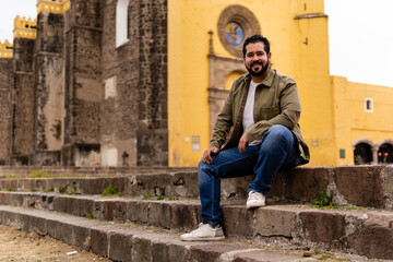 Fototapeta na wymiar Young mexican bearded guy with a jacket sitting on stone steps in a franciscan convent in Mexico. Full body portrait.