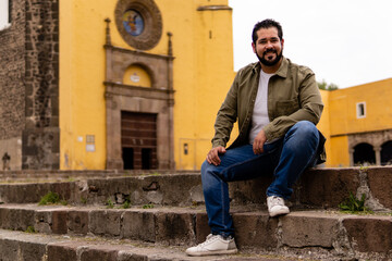 Young mexican bearded guy with a jacket sitting on stone steps in a franciscan convent in Mexico. Full body portrait.