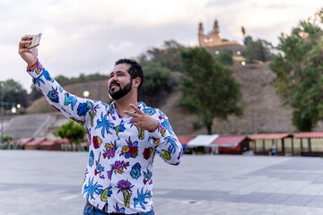 Young bearded man wearing a colorful flower shirt taking a selfie and smiling in front of the...