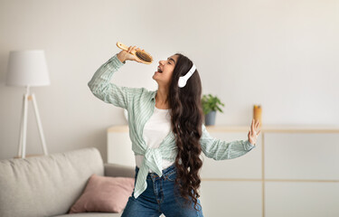 Cheerful Indian woman with hairbrush as mic singing her favorite song, wearing headphones and dancing at home