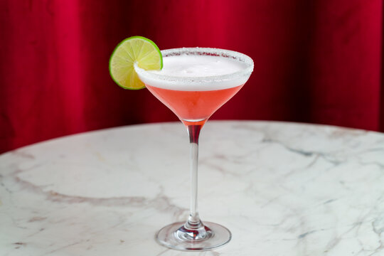 Cosmopolitan Cocktail With Lime And Salted Rim On Marble Table And Red Curtain Behind