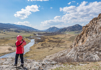 Naklejka premium a little girl, a child in an autumn warm jacket stands and takes pictures with a camera of a landscape with a river, standing on a stone of a high mountain