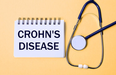 On a beige background, a stethoscope and a white notepad with the inscription CROHN IS DISEASE....