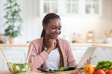 Happy african female checking recipe on digital tablet while cooking in kitchen