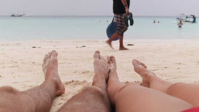 POV of Feet of a young couple of men and women lying on a tropical sandy beach near Ocean. Male and female legs sunbathe and relax on the exotic resort. Romantic vacation for couple in love. Zanzibar