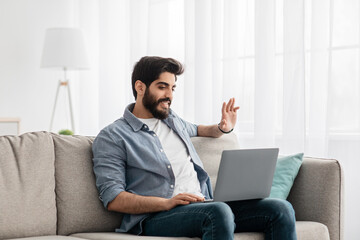 Distant communication. Arab man video calling on laptop computer, waving hand to screen while...