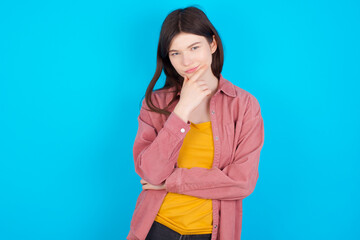 Thoughtful smiling young beautiful Caucasian woman wearing pink jacket over blue wall keeps hand under chin, looks directly at camera, listens something with interest. Youth concept.