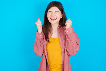 young beautiful Caucasian woman wearing pink jacket over blue wall rejoicing his success and victory clenching fists with joy being happy to achieve aim and goals. Positive emotions, feelings.