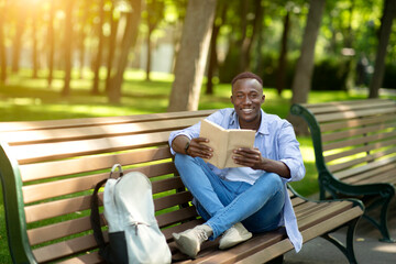 Fototapeta na wymiar Funky African American guy reading fascinating book on bench at urban park, empty space