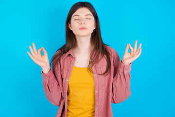 young beautiful Caucasian woman wearing pink jacket over blue wall doing yoga, keeping eyes closed, holding fingers in mudra gesture. Meditation, religion and spiritual practices.