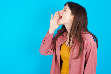 young beautiful Caucasian woman wearing pink jacket over blue wall profile view, looking happy and excited, shouting and calling to copy space.