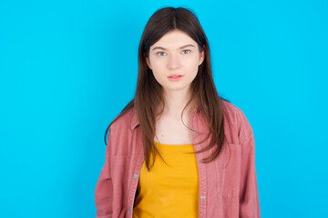 Joyful young beautiful Caucasian woman wearing pink jacket over blue wall looking to the camera, thinking about something. Both arms down, neutral facial expression.