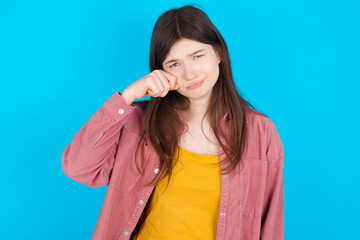 Unhappy young beautiful Caucasian woman wearing pink jacket over blue wall crying while posing at camera whipping tears with hand.