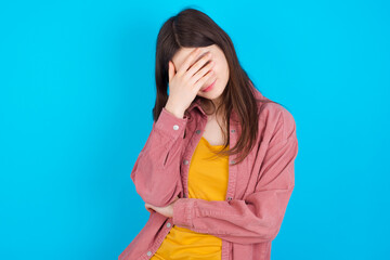 young beautiful Caucasian woman wearing pink jacket over blue wall making facepalm gesture while smiling amazed with stupid situation.