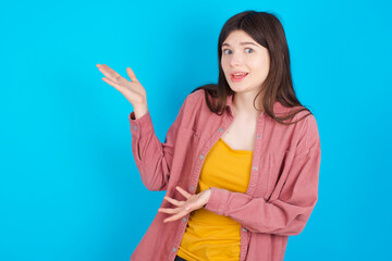 young beautiful Caucasian woman wearing pink jacket over blue wall pointing aside with both hands showing something strange and saying: I don't know what is this. Advertisement concept. - obrazy, fototapety, plakaty