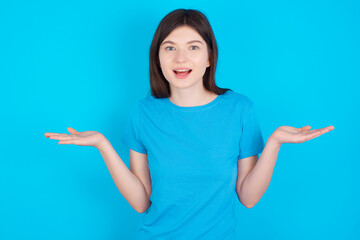 So what? Portrait of arrogant young beautiful Caucasian woman wearing blue T-shirt over blue wall shrugging hands sideways smiling gasping indifferent, telling something obvious.