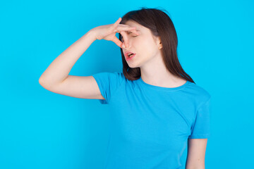 Displeased young beautiful Caucasian woman wearing blue T-shirt over blue wall plugs nose as smells...