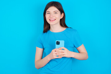 Smiling young beautiful Caucasian woman wearing blue T-shirt over blue wall friendly and happily holding mobile phone taking selfie in mirror.