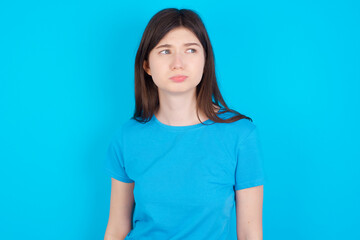 Dissatisfied young beautiful Caucasian woman wearing blue T-shirt over blue wall purses lips and has unhappy expression looks away stands offended. Depressed frustrated model.