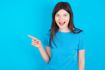 young beautiful Caucasian woman wearing blue T-shirt over blue wall points aside on copy blank space. People promotion and advertising concept