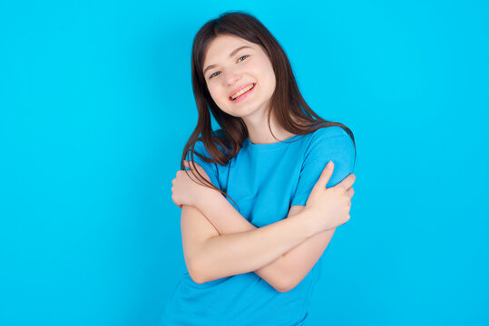 Charming pleased young beautiful Caucasian woman wearing blue T-shirt over blue wall embraces own body, pleasantly feels comfortable poses. Tenderness and self esteem concept