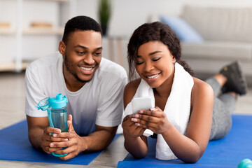Fit African American couple lying on yoga mats with cellphone after domestic workout, checking new sports videos online