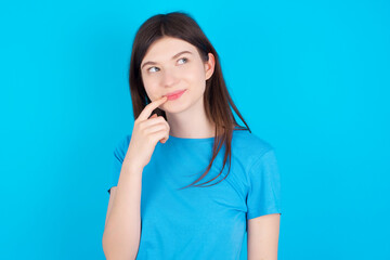 Lovely dreamy young beautiful Caucasian woman wearing blue T-shirt over blue wall keeps finger near lips looks aside copy space.