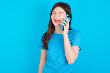 Overemotive happy young beautiful Caucasian woman wearing blue T-shirt over blue wall laughs out positively hears funny story from friend during telephone conversation