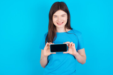 Fototapeta premium Cheerful cheery content young beautiful Caucasian woman wearing blue T-shirt over blue wall holding in hands device hobby smm post blog