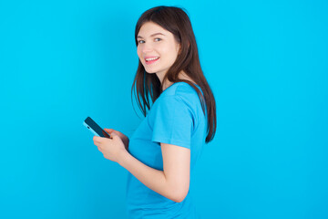 Fototapeta premium Rear view photo portrait of young beautiful Caucasian woman wearing blue T-shirt over blue wall using smartphone smiling