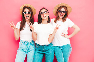 Three young beautiful smiling hipster female in trendy same summer white t-shirt and jeans clothes. Sexy carefree women posing near pink wall in studio. Positive models showing peace sign