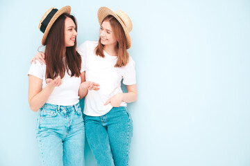 Obraz na płótnie Canvas Two young beautiful smiling hipster female in trendy summer white t-shirt and jeans clothes.Sexy carefree women posing near light blue wall in studio.Positive models in hats communicating