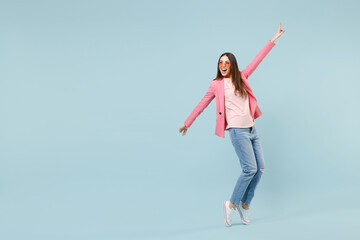 Full length young stylish woman 20s in pastel pink clothes glasses standing on toes dancing leaning back with outstretched hands isolated on blue background studio portrait. People lifestyle concept.