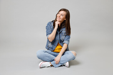 Full length young happy dreamful caucasian woman in casual denim jacket yellow tshirt sitting cross-legged prop up chin look aside isolated on grey background studio portrait People lifestyle concept