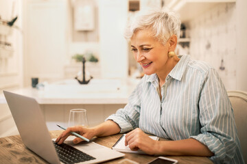 Attractive middle aged female freelancer in striped shirt sitting at home in front of open laptop,...